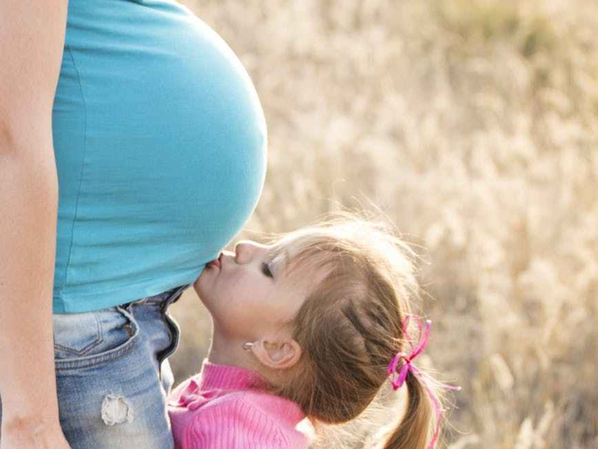 girl in pink sweater and grey jeans kissing tummy of pregnant woman in blue shirt and blue denim jeans